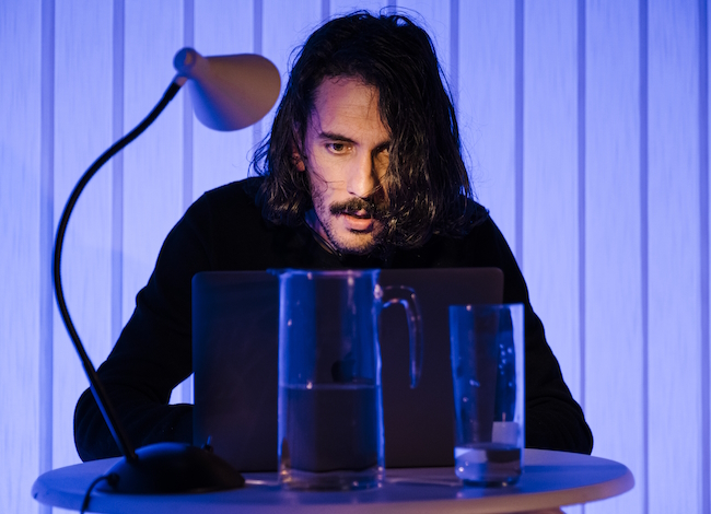 Il y a une table avec une carafe d'eau, un verre, une lampe et un homme regarde cette table
